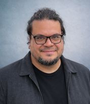 Retrato: sonríe a cámara, con gafas de pasta negras, barba y chaqueta oscura, ante un fondo gris azulado.