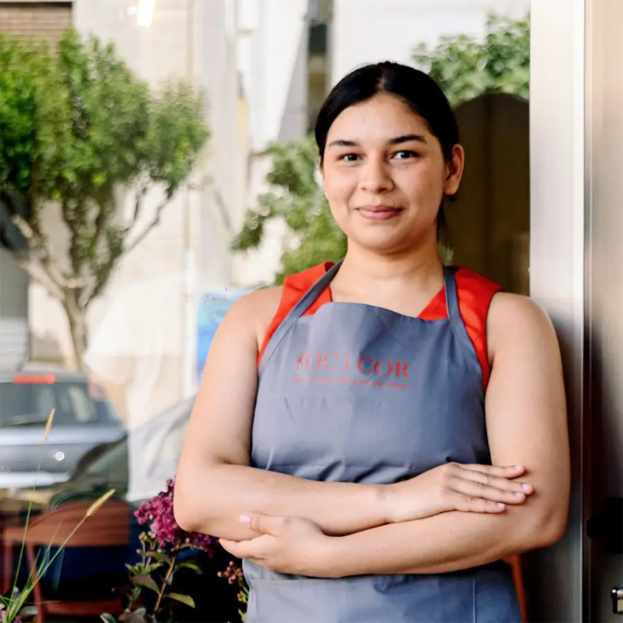 Una mujer del equipo, con el delantal de Foc i Cor y los brazos cruzados, sonríe apoyada en la puerta del restaurante.